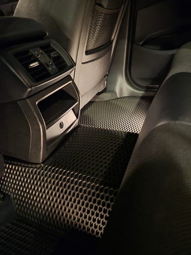Recenzent pokazuje czarne dywaniki samochodowe o strukturze plastra miodu, idealnie dopasowane do podłogi w tylnej części auta.