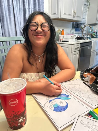 Customer smiling while wearing the CHAIN OF REPUTATION pearl necklace at a table with a coloring book.