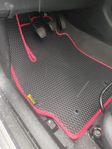 Recenzent pokazuje czarny dywanik samochodowy z czerwoną lamówką, zamontowany po stronie kierowcy.