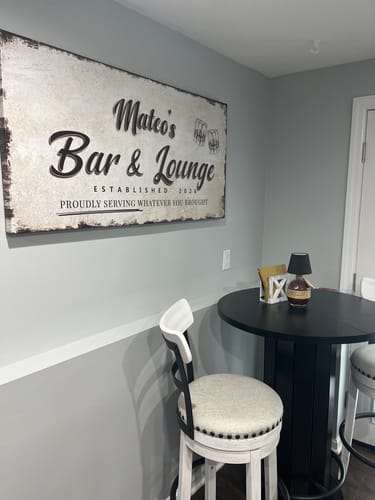 A customer's large, rustic Personalized Basement Bar Sign hanging on a gray wall in their home bar area.