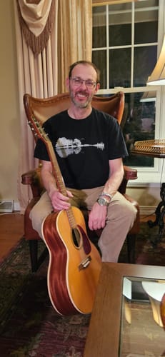 Customer wearing the black Forest Guitar T-Shirt while sitting in a chair and holding an acoustic guitar.