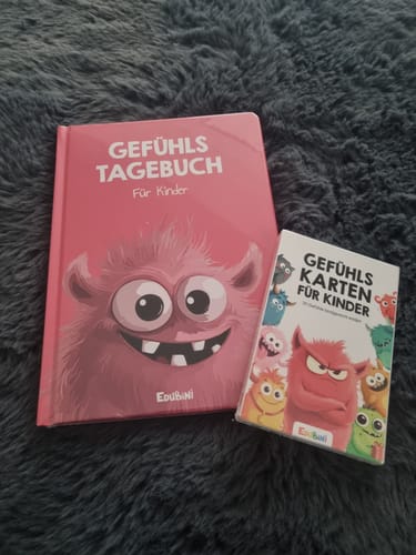 Ein Kunde zeigt das rosa Gefühlstagebuch für Kinder mit den dazugehörigen Gefühlskarten auf einem Teppich.