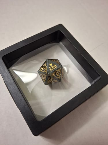 Customer's personalized dark D20 dice with gold engraving in a display case. The '20' face has the initials 'S & A' and a date.