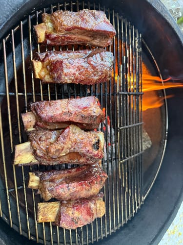 Cliente cocinando seis costillas Short Rib en una parrilla redonda con fuego visible.
