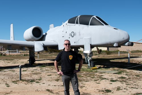Customer wearing the black heavyweight t-shirt with a small chest logo, standing in front of a military airplane.