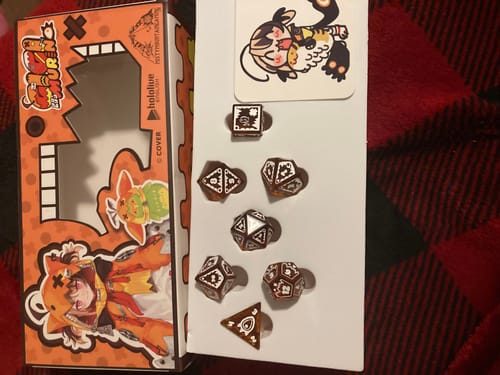 Customer's Gigi Murin Glass Dice Set and sticker displayed next to the colorful product packaging on a blanket.
