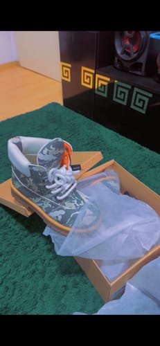 A customer's Camo Print Ankle Boot with a tan sole and white laces, shown in its open box on a green rug.