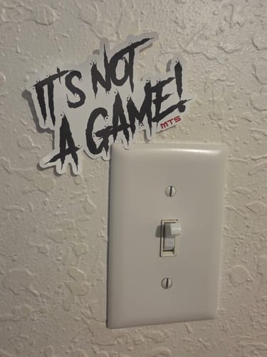 Customer's black and white "IT'S NOT A GAME!" sticker from the TF Premium Sticker Pack on a textured wall.