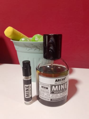 Il recensore mostra il flacone di profumo ARCHÉ e un campioncino MINE su un tavolo.
