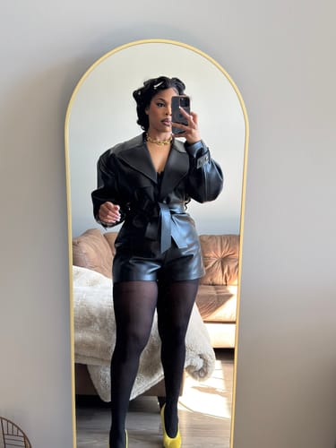 Reviewer in a mirror selfie wearing the black Vegan Leather Trench Romper with pantyhose and yellow heels.