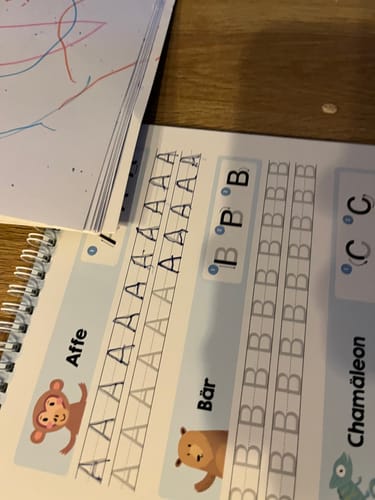 Kunde zeigt ein aufgeschlagenes Edubini Magisches Übungsheft mit nachgezeichneten Buchstaben auf einer Holzoberfläche.