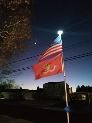 Customer's United States Marines Flag waving below an American flag on a lit flagpole at dusk.
