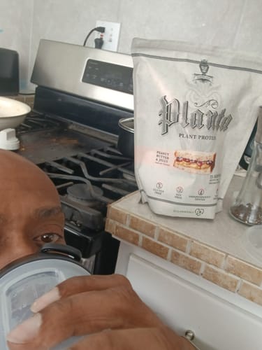 Close-up of a reviewer drinking from a shaker with a bag of Planta plant protein on a kitchen counter.