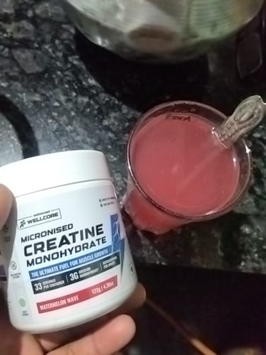 Reviewer holds the Wellcore Creatine Monohydrate container next to a glass of the reddish, mixed drink.