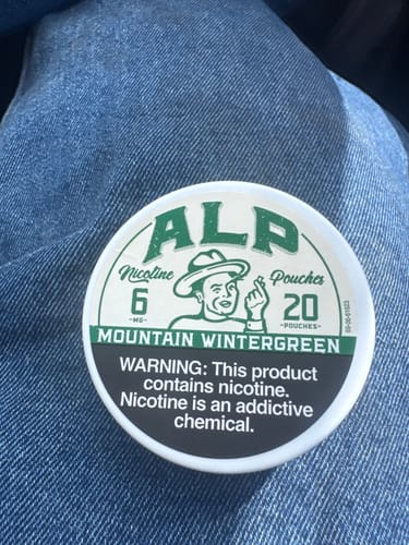 Customer's white and green tin of ALP Mountain Wintergreen Nicotine Pouches resting on their denim-clad lap.
