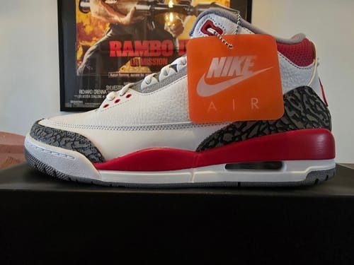 Klant toont de Air Jordan 3 OG Fire Red schoen in zijaanzicht op de doos, met een oranje Nike-tag.