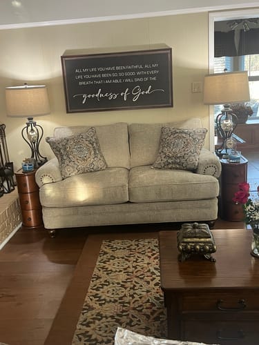 Customer's beautiful Goodness Of God Faith Sign II hanging above a loveseat in a warm, welcoming living room.