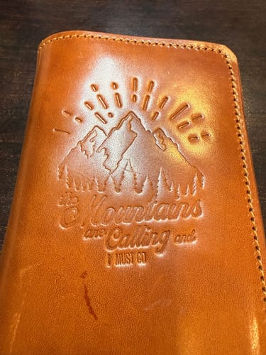 Close-up of a customer's brown leather Field Notes Wallet, showing the embossed 'Mountains are Calling' design on the cover.
