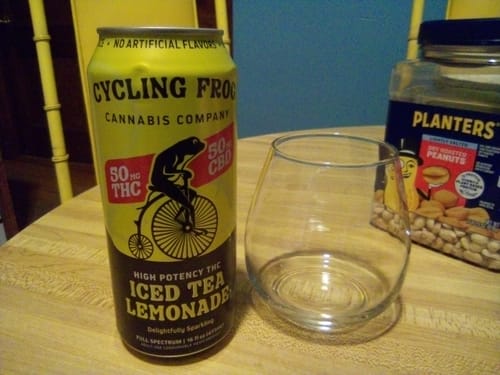 Reviewer's yellow can of Sparkling THC Iced Tea Lemonade on a wooden table next to an empty stemless glass.