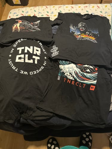 Reviewer showing four different graphic t-shirts from their TCG91 MYSTERY CASH BOX laid out on a bed.