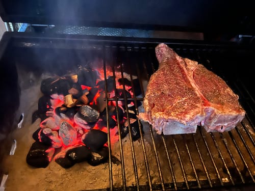 Customer's seasoned USDA Choice Porterhouse Steak on a grill next to glowing charcoal before cooking.