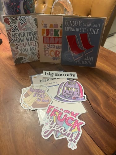 Customer's 'Congrats On No Longer Having To Give A F*ck' card displayed with other fun cards and stickers on a table.