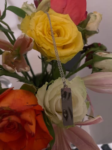 Ein Kunde präsentiert die Halskette mit Augen-Anhänger vor einem bunten Blumenstrauß.