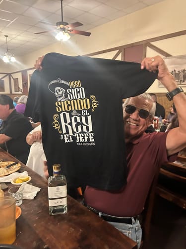 Reviewer's photo of a smiling person holding up the black Sigo Siendo El Rey T-Shirt.