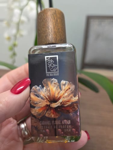 Customer's hand holding the Caramel Floral Affair perfume bottle, showing its floral label and wooden cap.