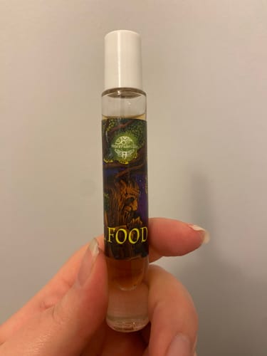 Reviewer holding a small glass rollerball of Troll Food perfume, showing the amber liquid and illustrated label against a plain wall.
