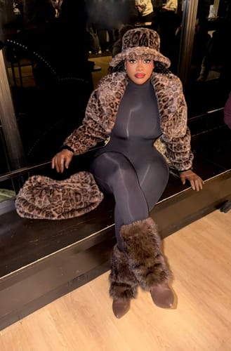 Reviewer sitting indoors wearing a black catsuit with a matching leopard print hat and bag.
