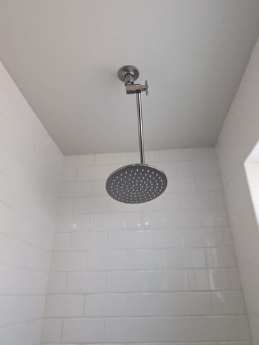 Reviewer's adjustable rain shower head and arm installed on the ceiling of a white tiled shower.