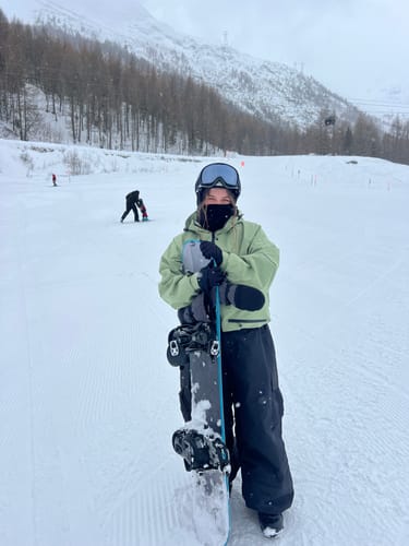 Reviewer wearing a light green snow jacket and holding a snowboard on a snowy slope.