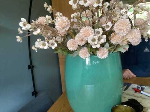 Beoordelaar toont het crèmekleurige Wax Arrangement in een grote blauwgroene vaas.