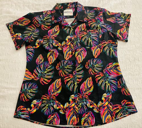 Reviewer's Vacation Steve shirt laid flat, showing its vibrant tropical leaf pattern on a black background.