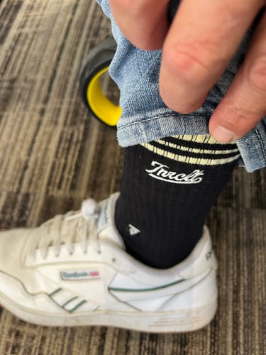 Close-up of a customer lifting their jean cuff to show the black SCRIPT CREW SOCKS with a white logo.