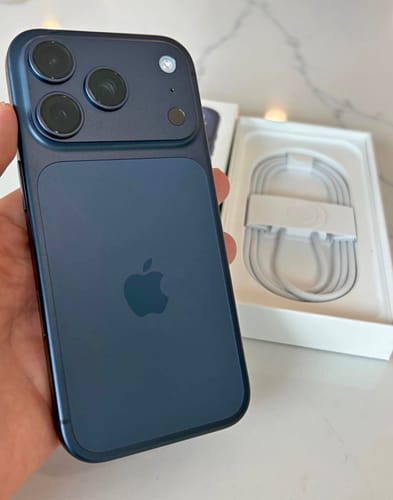 Cliente sosteniendo un IPHONE 17 PRO MAX azul oscuro en la mano, con la caja abierta al fondo.