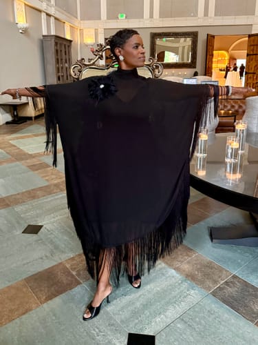 Reviewer in a black tasseled maxi dress with arms out, showing the loose batwing sleeves indoors.
