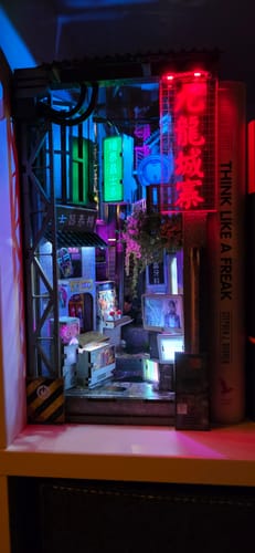 Customer's assembled Kowloon Walled City book nook kit glowing with vibrant neon lights on a bookshelf.
