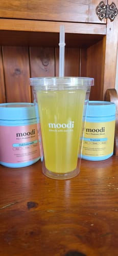 Customer's yellow Tropican Pre + Probiotic Blend prepared in a clear tumbler, with the product tubs on a wooden shelf.
