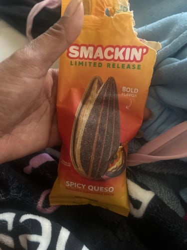 A person's hand holding an opened orange bag of Smackin' limited release Spicy Queso sunflower seeds.
