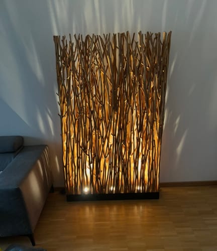 Kunde zeigt den beleuchteten SKOV Holz-Raumteiler, der in einem Wohnzimmer neben einem Sofa für warmes Licht sorgt.
