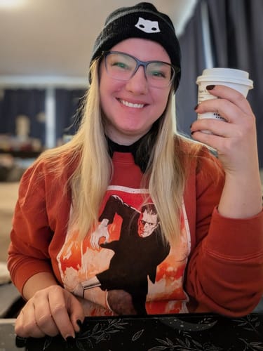 Reviewer smiling in a rust-red Frankenstein sweatshirt, styled with a black collar and holding a coffee cup.
