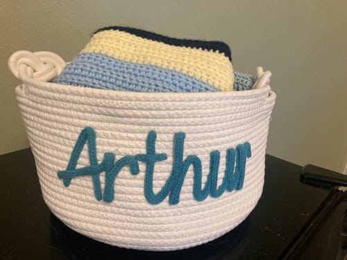 Reviewer's large, white rope basket personalized with 'Arthur' in teal yarn, holding a crocheted blanket.