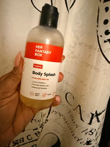 Reviewer holding a clear bottle of Yummy Body Splash body oil, showing the light-yellow liquid inside.