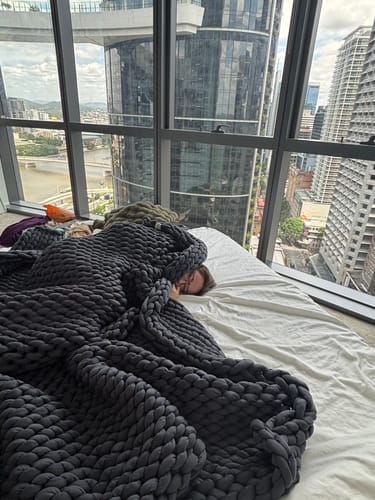 Reviewer relaxing in bed under a dark gray chunky Knitted Weighted Blanket in a high-rise room.