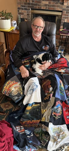 Customer in a recliner with a puppy, covered by their large Premium T Shirt Quilt made from various graphic t-shirts.