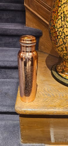 Customer's shiny, engraved copper water bottle standing on a wooden surface next to stairs.