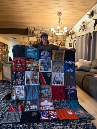 Reviewer proudly holding up a large Premium T Shirt Quilt made from many different shirts in a living room.