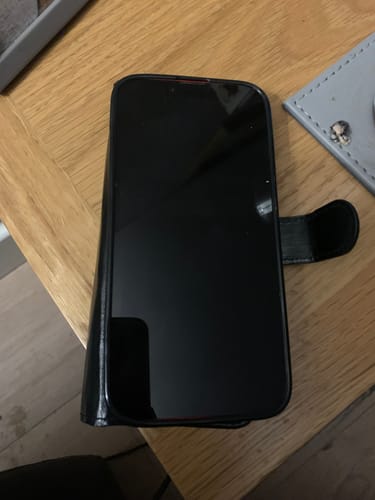 Customer's refurbished Apple iPhone 14 in a black wallet case, resting on a wooden table with the screen off.